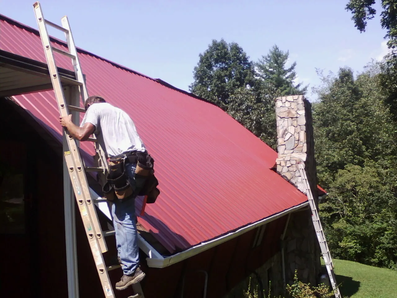 Roofer on a ladder beside a red metal roof and stone chimney — Soffit & Fascia Repair by Roofing Brothers in Goshen